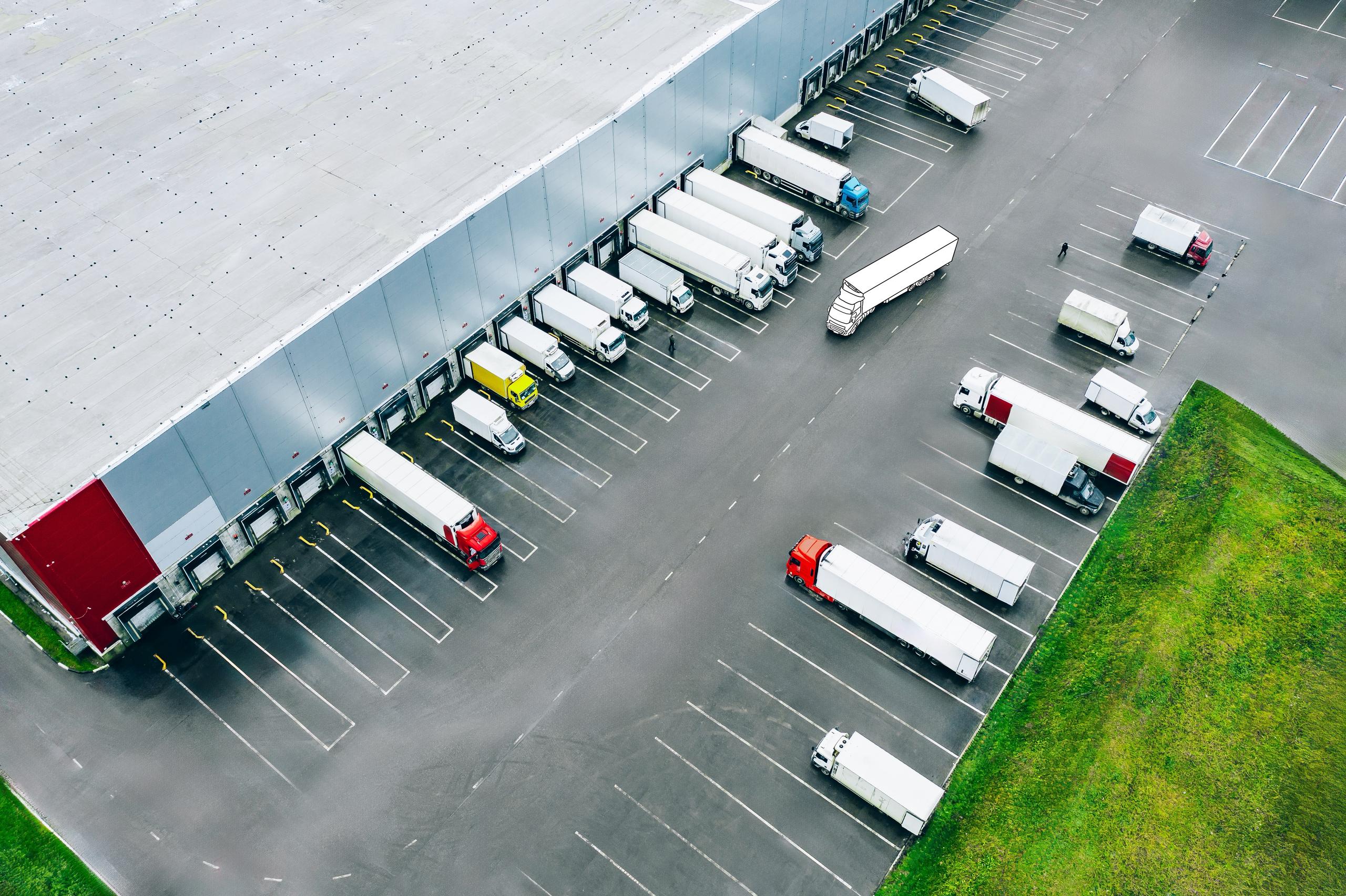 Luftaufnahme eines Logistikzentrums: An der langen roten und grauen Gebäudefassade mit zahlreichen Laderampen stehen mehrere LKW und Sattelanhänger zum Be- und Entladen. Weitere Fahrzeuge parken auf dem angrenzenden Parkplatz, rechts im Bild eine Rasenfläche.